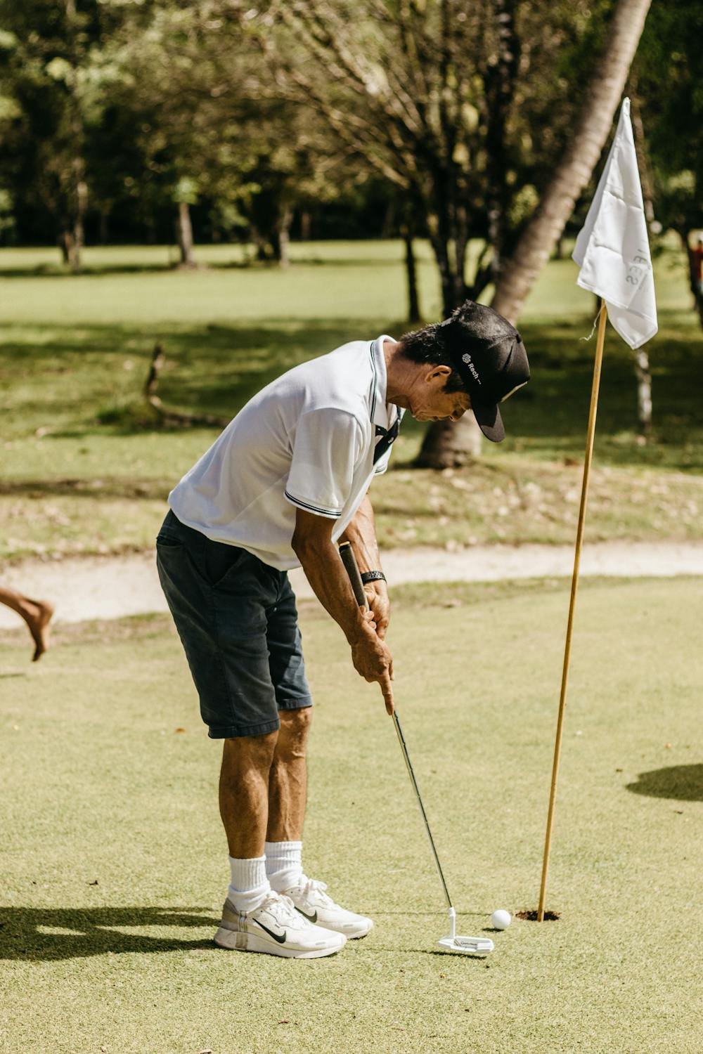 A man swings a golf club on a lush green course, focusing intently on his shot. The scene captures the moment of impact as the club makes contact with the ball, with a clear blue sky and well-maintained fairway in the background.